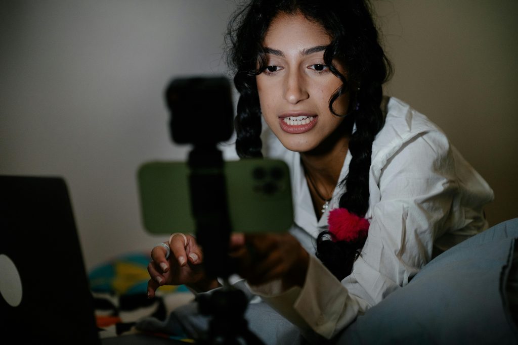 A young woman with braided hair records a video using her smartphone and laptop indoors.